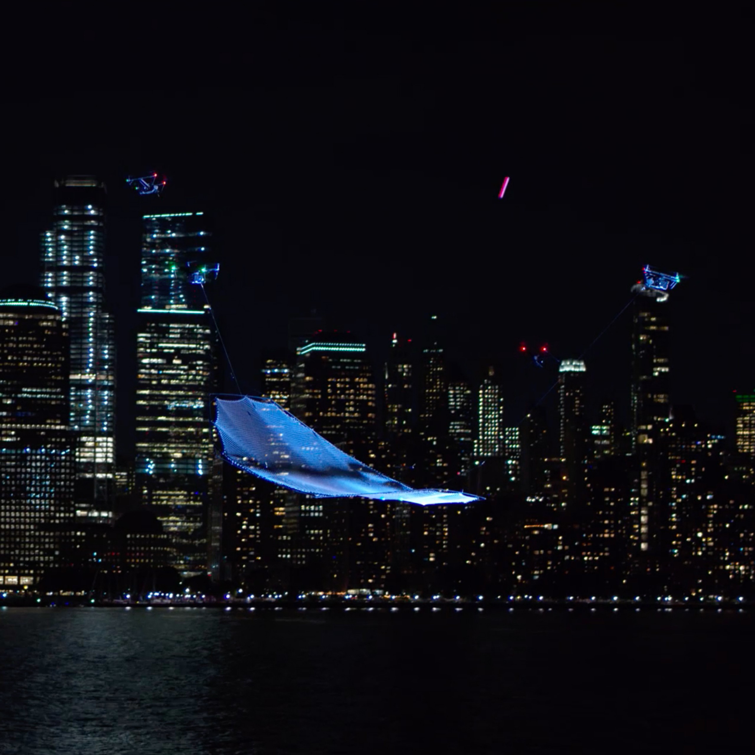 Vista de una ciudad de noche, con una red sostenida en el cielo por tres drones.